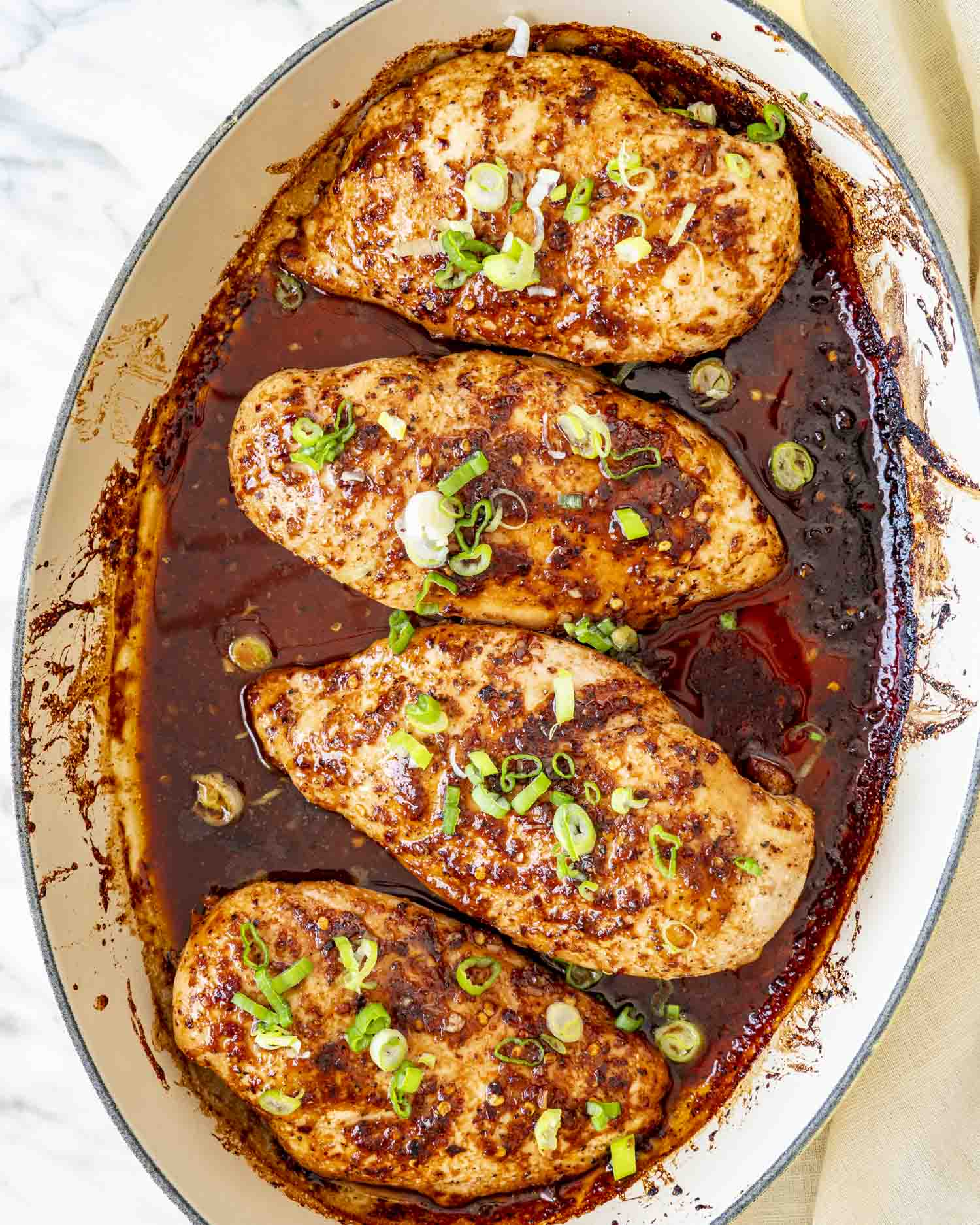 Overhead view of four baked firecracker chicken breasts in skillet, surrounded by caramelized sauce and scattered chopped green onions.