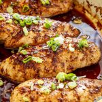 Several firecracker chicken breasts nestled in spicy sauce, glistening with baked glaze and sprinkled generously with fresh scallions.