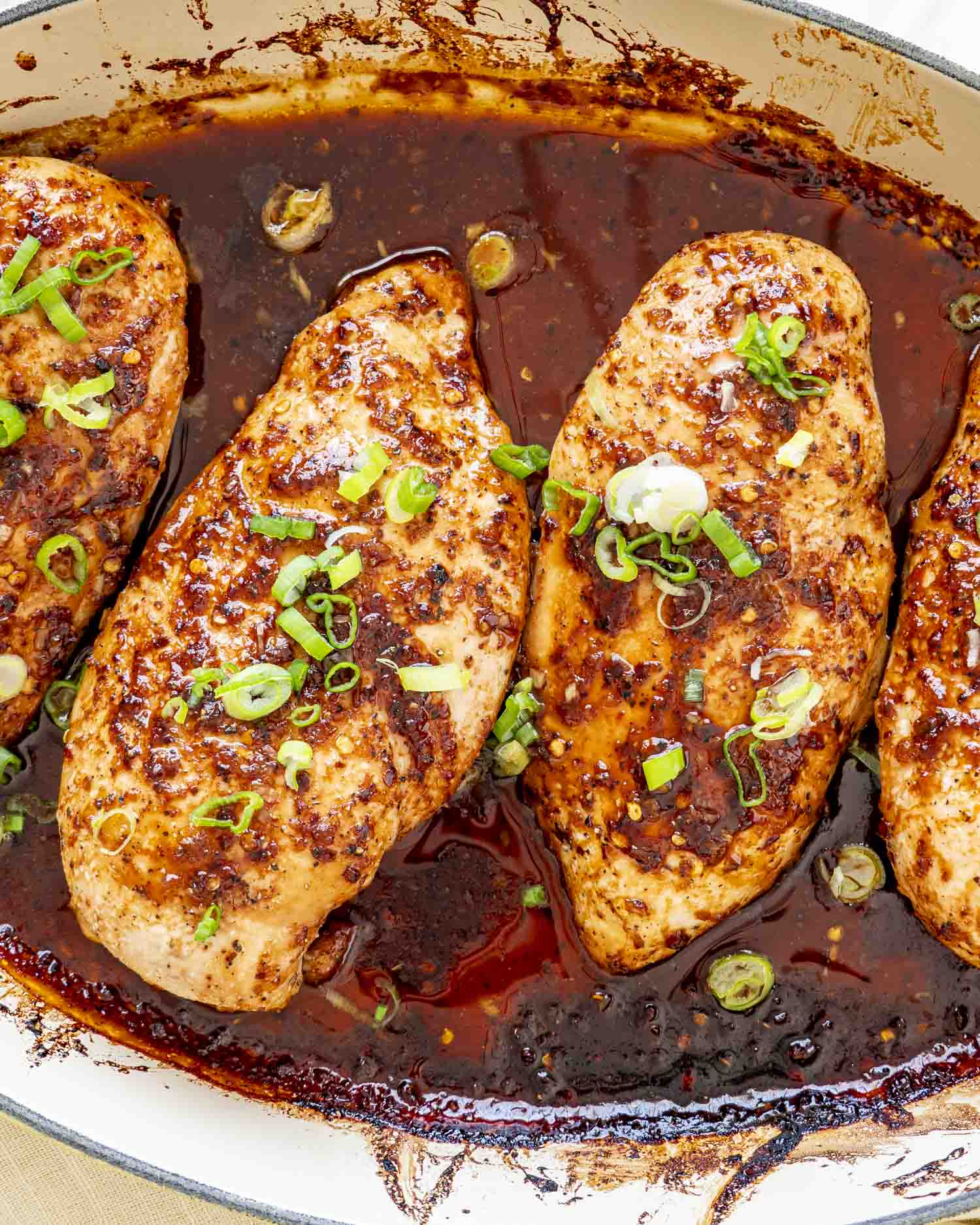 Close-up of baked firecracker chicken breasts in a skillet, coated in glossy sweet-spicy sauce and topped with scallions.