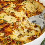 Close-up of loaded scalloped potatoes in a white dish, topped with cheese and bacon, with a serving spoon.