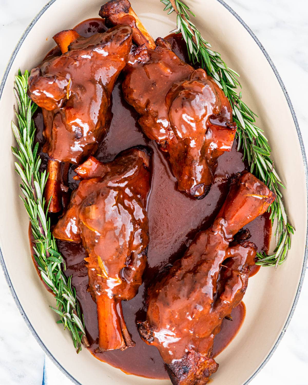 overhead shot of braised lamb shanks with sauce in a platter garnished with fresh rosemary