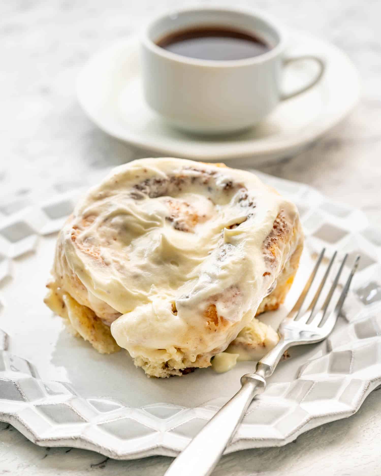 a cinnabon cinnamon roll on a white dessert plate with a cup of coffee in the background.
