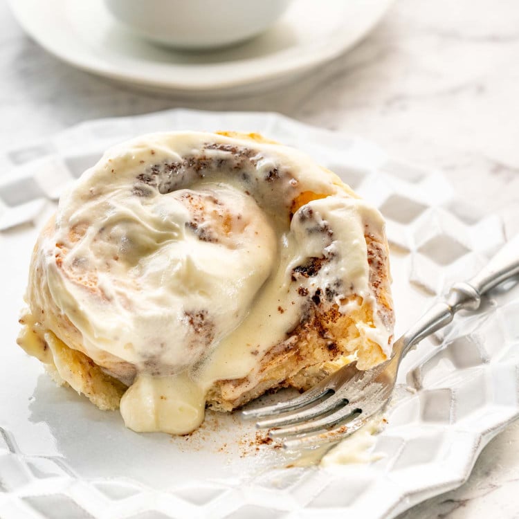 a cinnabon cinnamon roll on a white dessert plate with a cup of coffee in the background.