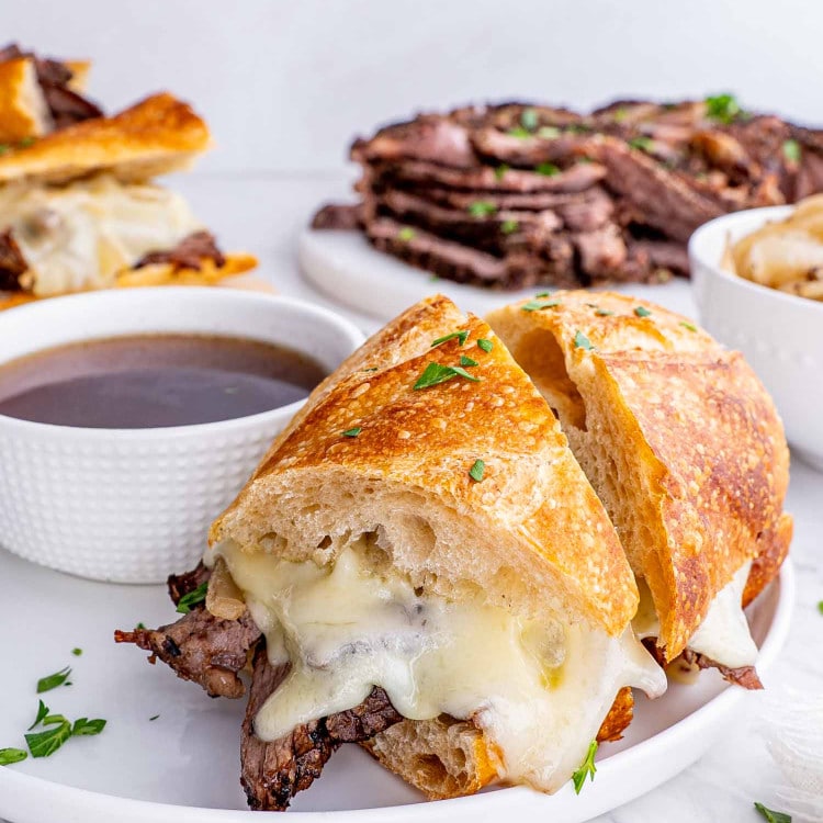 a french dip sandwich cu in half on a white plate next to a bowl with au jus.