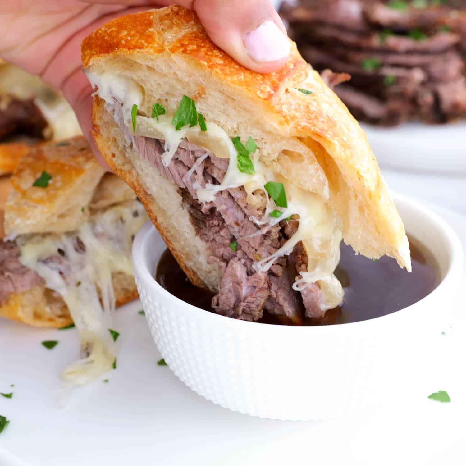a hand dipping a french dip sandwich in au jus.