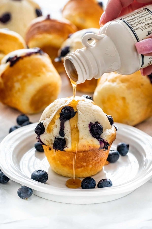a hand pouring syrup from a bottle over a blueberry pancake muffin on a plate