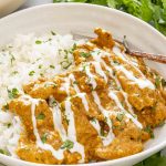 Close-up angled view of chicken korma with white rice, creamy sauce, yogurt drizzle, and cilantro garnish in a white bowl.