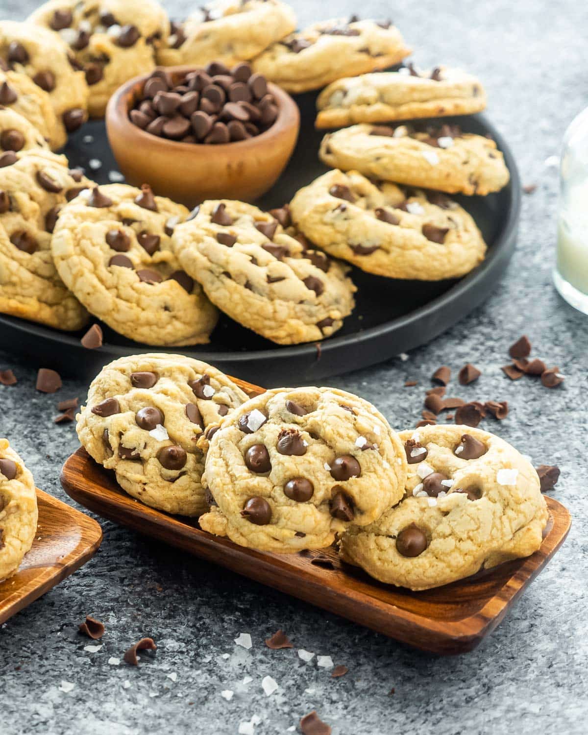 chocolate chip cookies on a wooden plate with a black plate filled with cookies in the back.