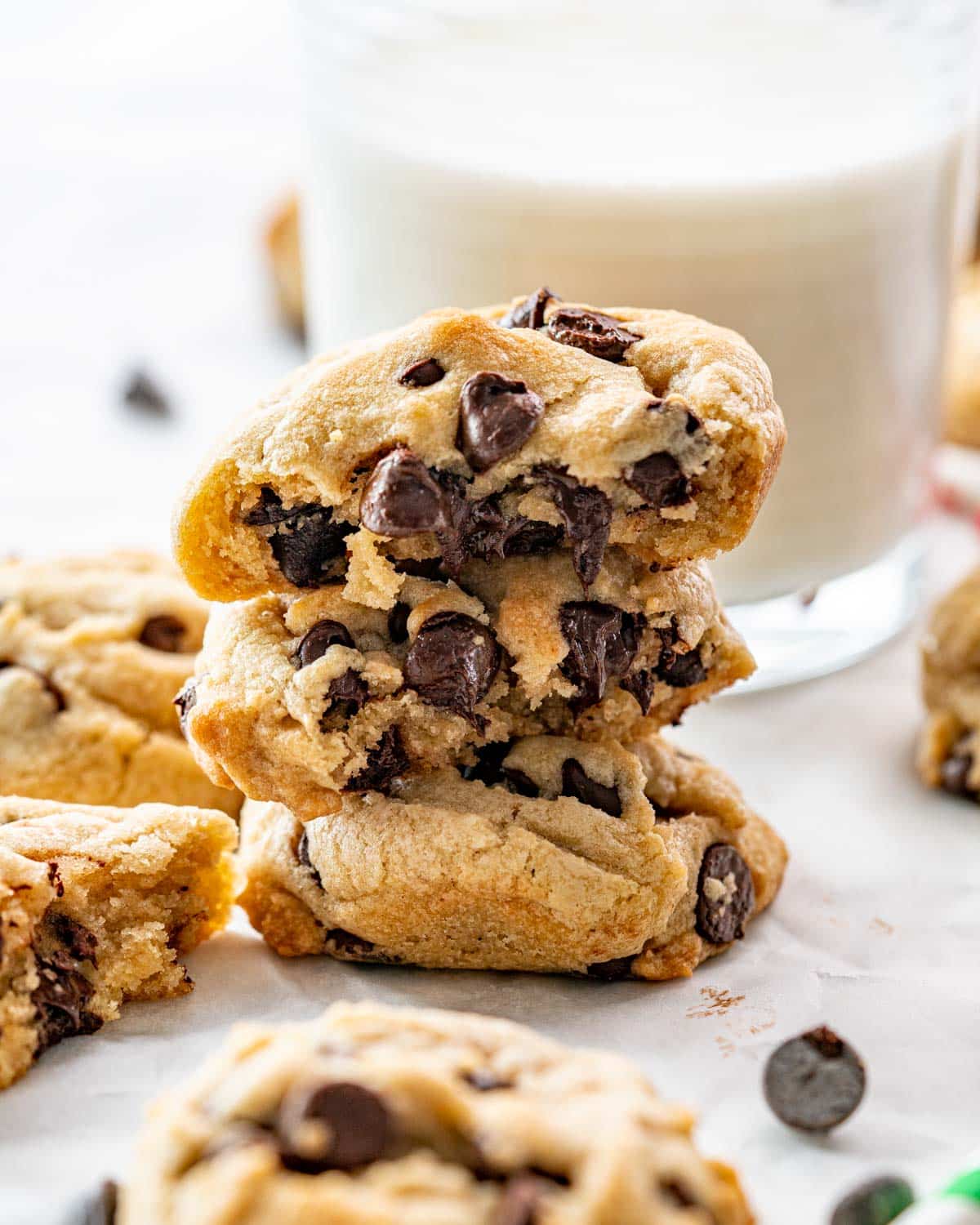 a stack of chocolate chip cookies with the top one broken in half and a milk glass in the back.