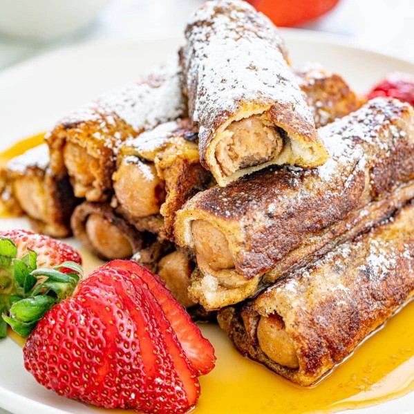 sausage french toast roll ups dusted with powdered sugar and topped with maple syrup - one has a bite taken from it, on a plate with whole sliced strawberries