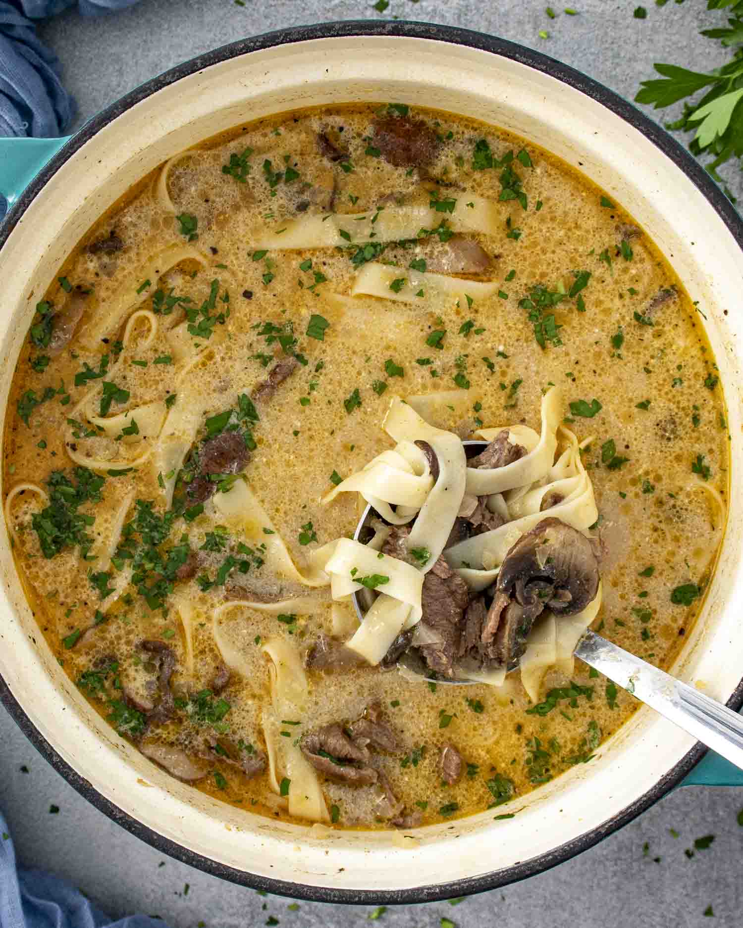 A close-up overhead shot of a Beef Stroganoff Soup in a large Dutch oven. The creamy, golden broth is filled with tender strips of beef, sliced mushrooms, and wide egg noodles, all garnished with freshly chopped parsley. A ladle is lifting a portion of the soup, showcasing the rich, comforting ingredients.