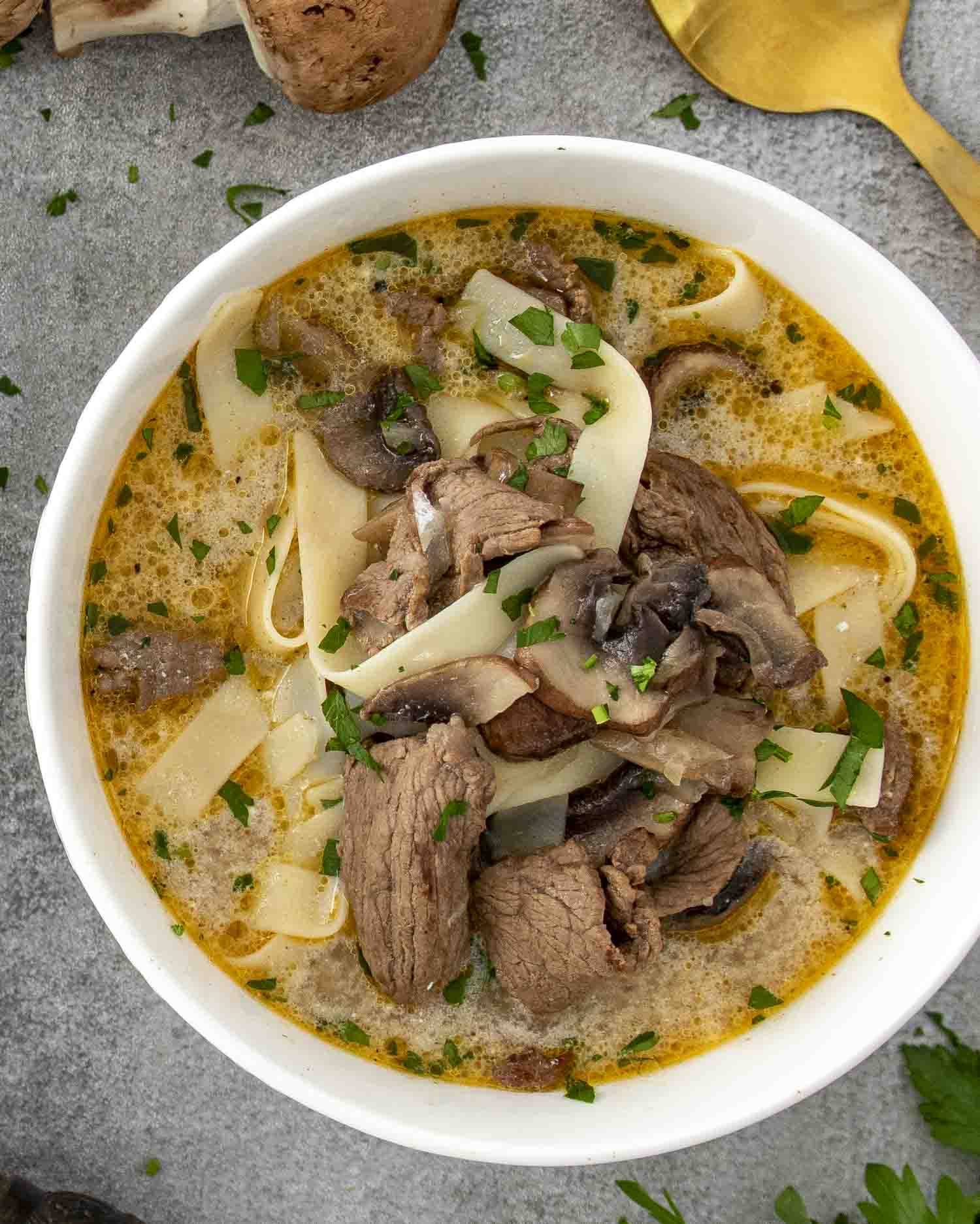 A close-up shot of a bowl of Beef Stroganoff Soup served in a textured white bowl on a matching plate. The creamy, golden broth is packed with tender slices of beef, wide egg noodles, and earthy mushrooms, all garnished with freshly chopped parsley.