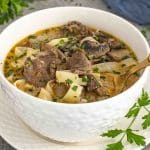 A close-up shot of a bowl of Beef Stroganoff Soup served in a textured white bowl on a matching plate. The creamy, golden broth is packed with tender slices of beef, wide egg noodles, and earthy mushrooms, all garnished with freshly chopped parsley.