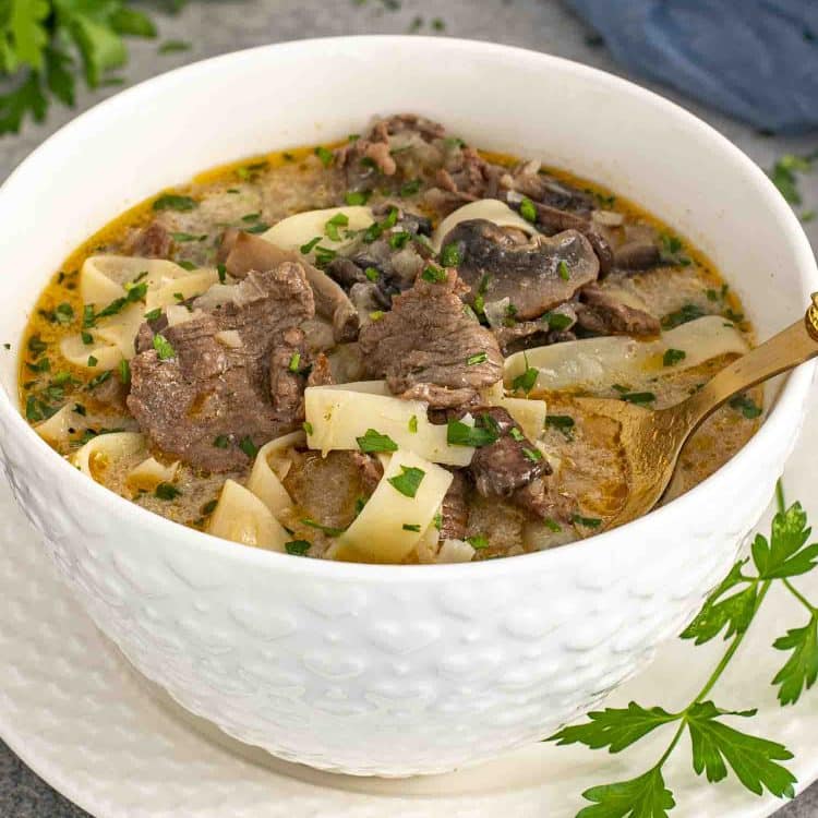 A close-up shot of a bowl of Beef Stroganoff Soup served in a textured white bowl on a matching plate. The creamy, golden broth is packed with tender slices of beef, wide egg noodles, and earthy mushrooms, all garnished with freshly chopped parsley.
