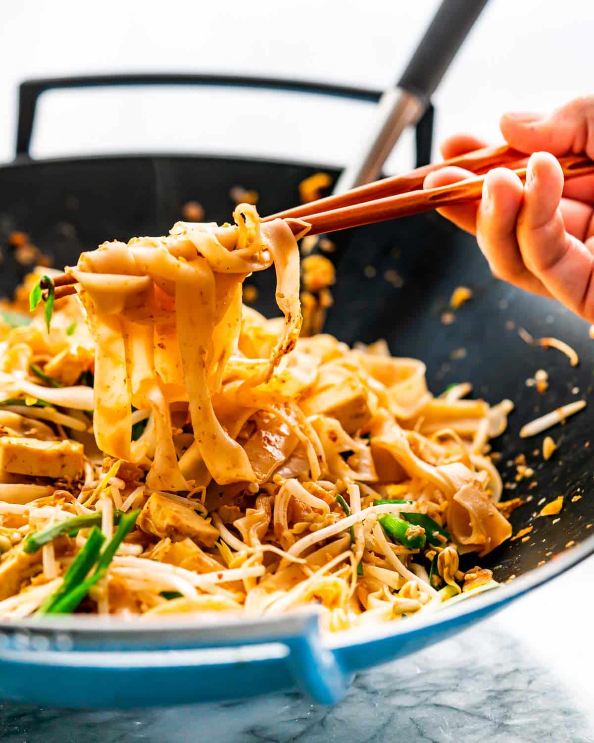 pad thai in a wok with a hand holding some noodles with chopsticks.