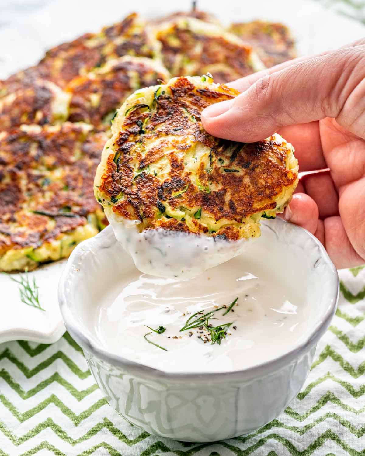 a hand dipping a zucchini fritter in a bowl with ranch dressing.