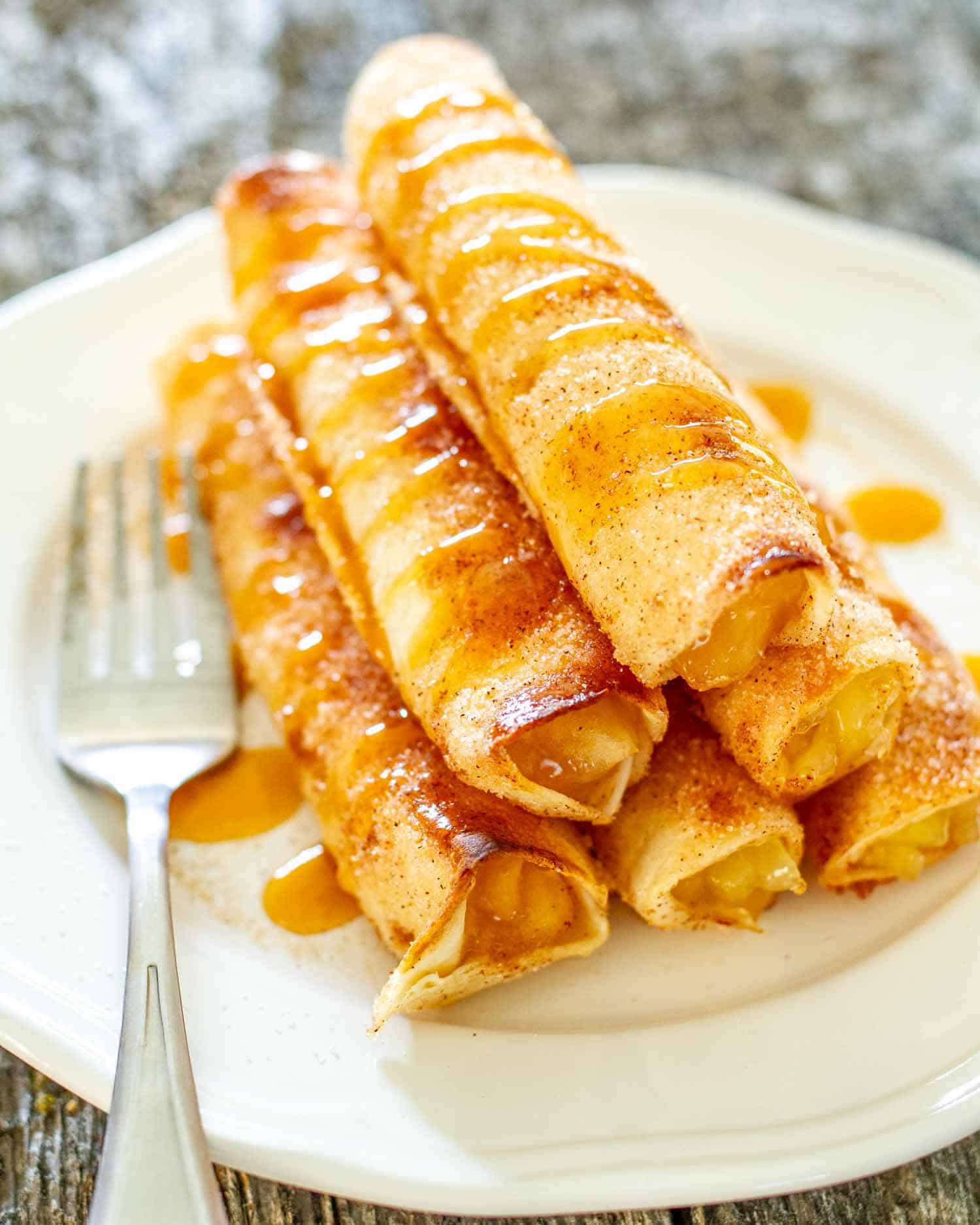 a stack of apple pie taquitos on a white plate drizzled with caramel sauce.