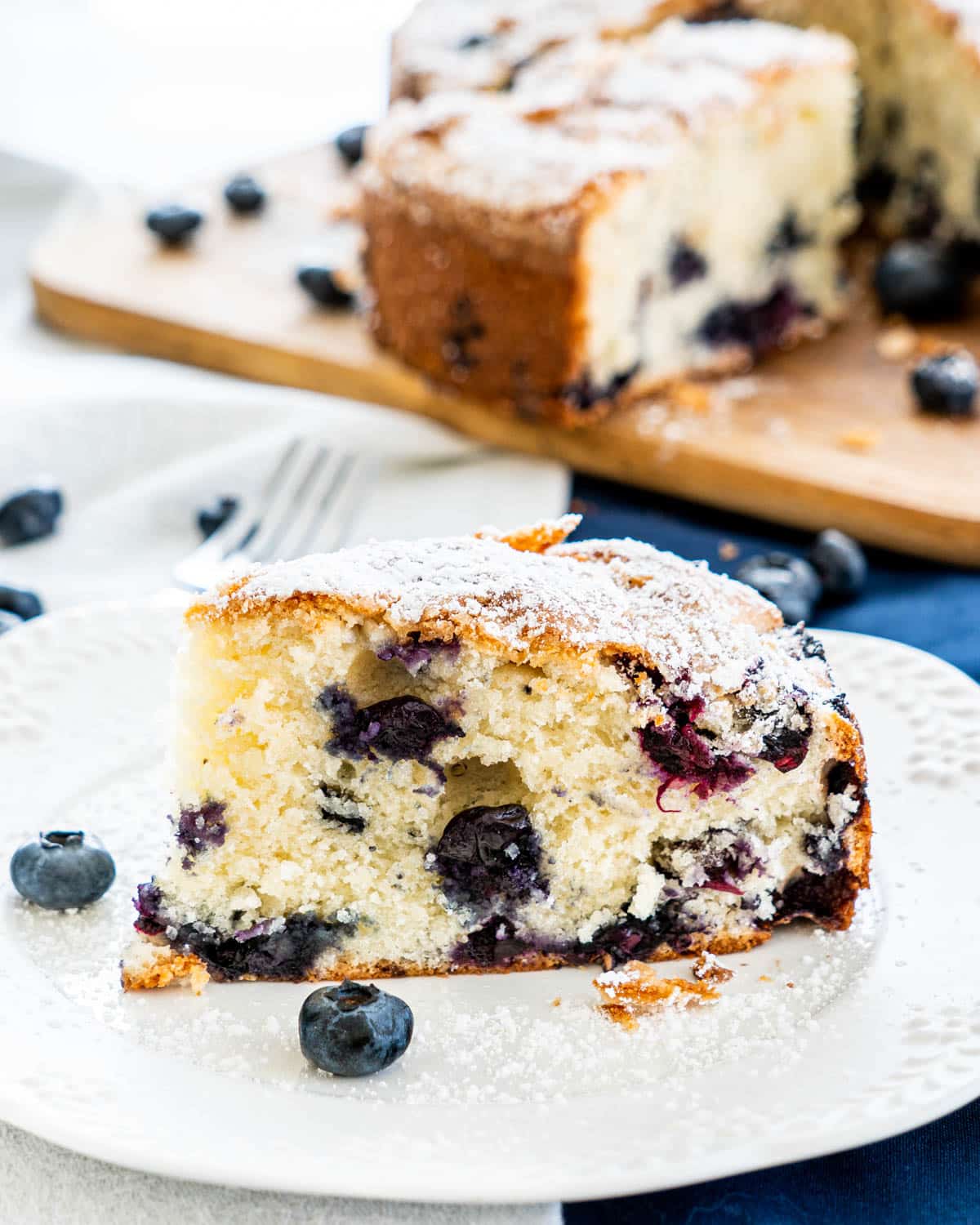 a slice of blueberry cake on a white plate with the rest of the cake in the background