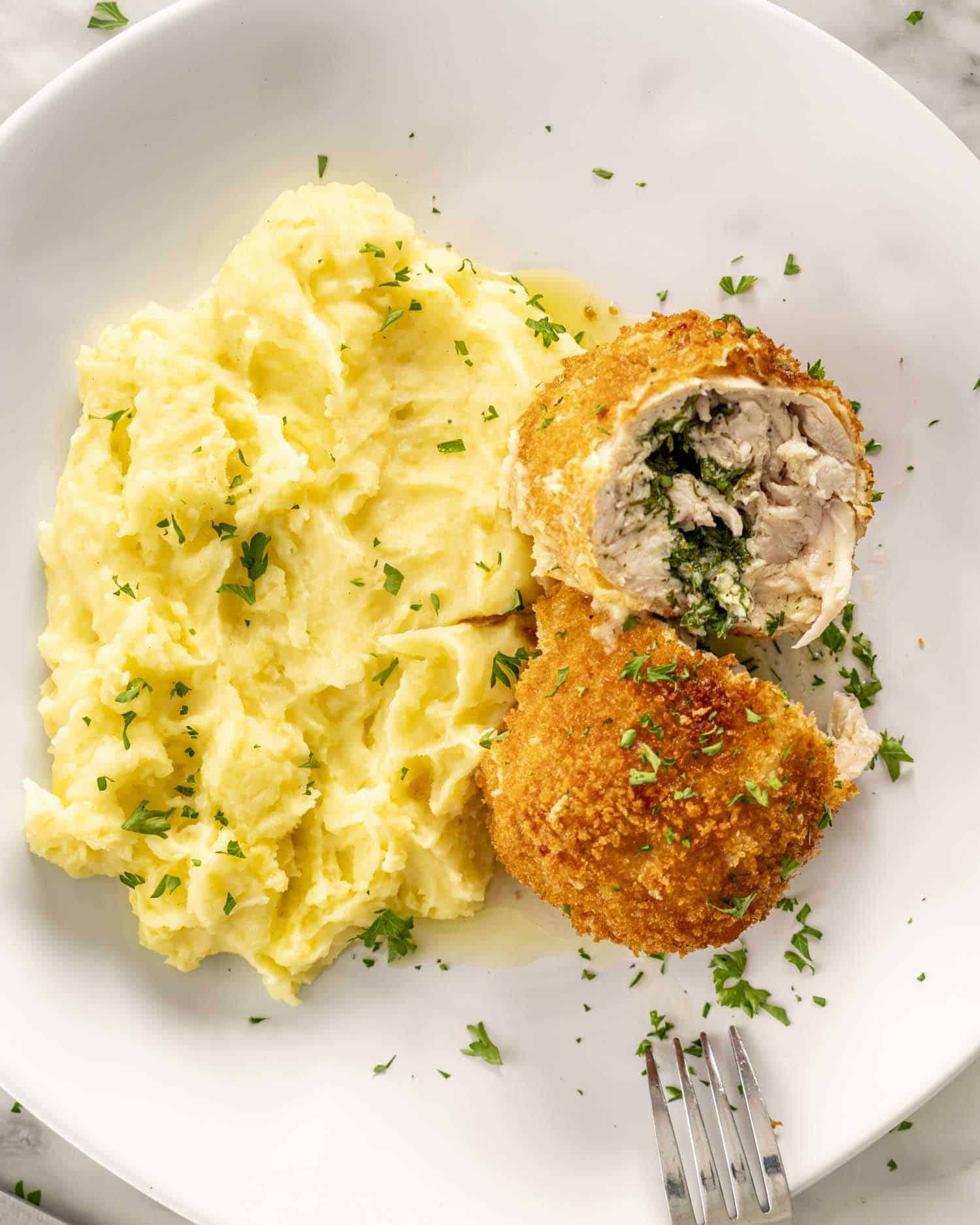Chicken Kiev Chicken Kiev on a white plate along mashed potatoes, cut in the middle with the herb garlic butter spilling from the center.