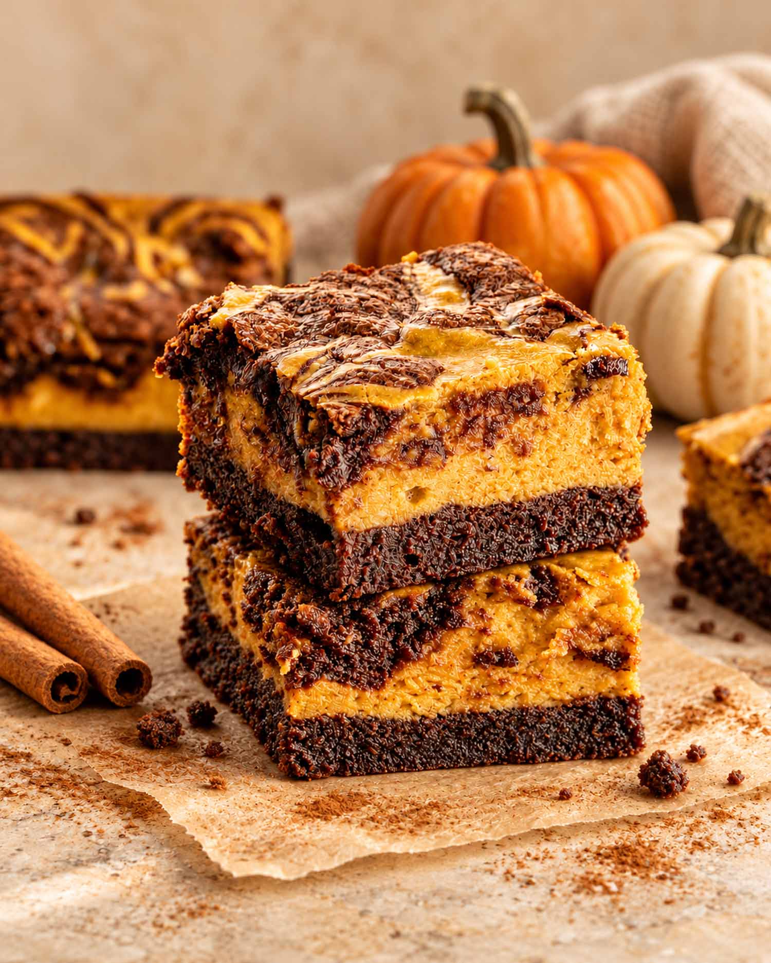 Two pumpkin cheesecake brownies stacked on parchment, showing chocolate bases and pumpkin layers, with small pumpkins and cinnamon sticks nearby.