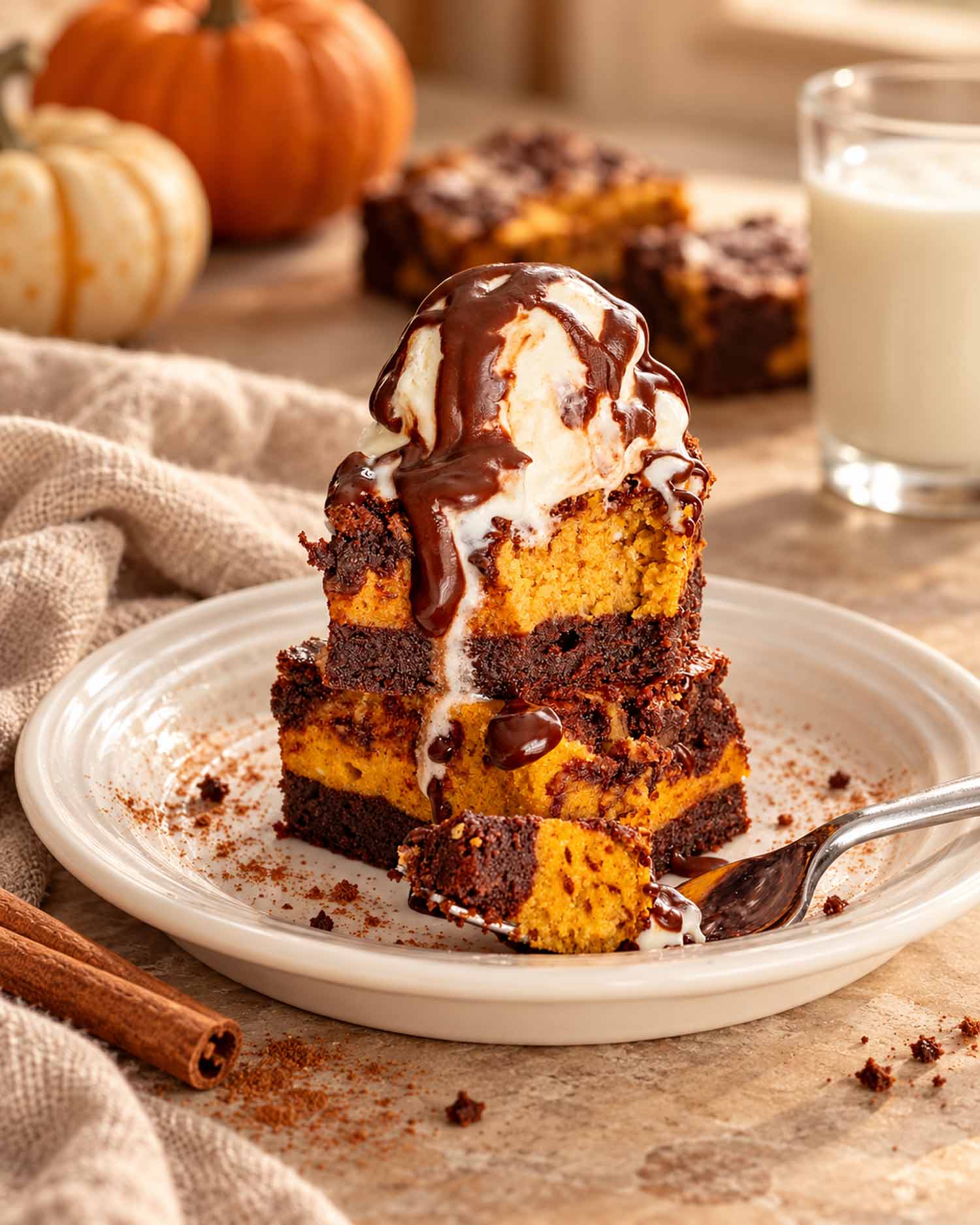 Stacked pumpkin cheesecake brownies topped with vanilla ice cream and chocolate sauce, served on a white plate beside a spoon.