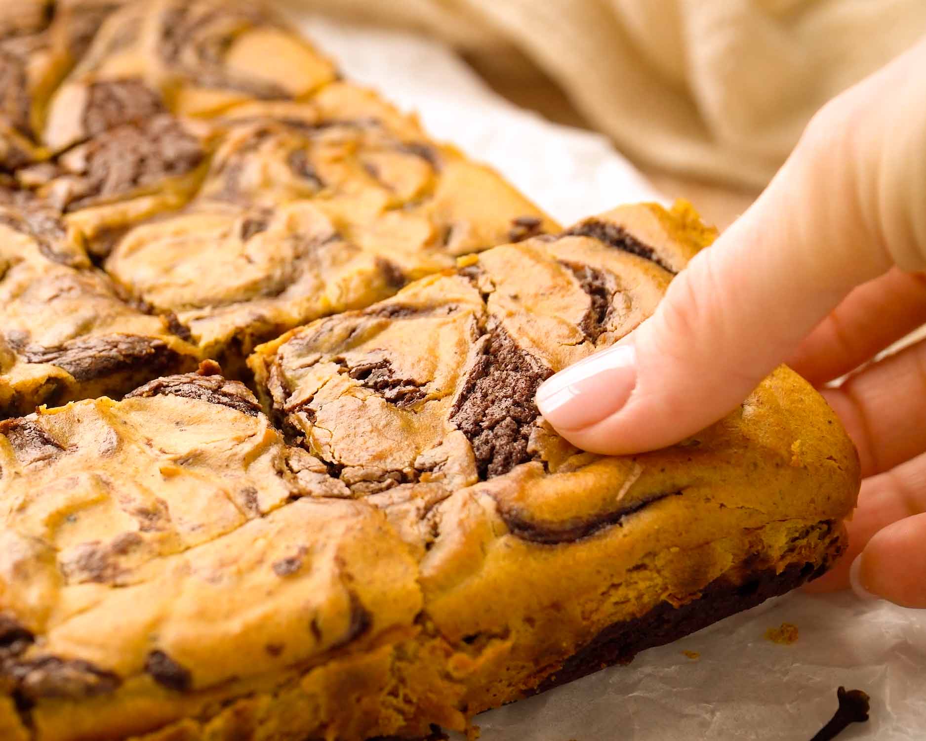 Hand lifting a pumpkin cheesecake brownie from a stack on a white plate, with pumpkins and cinnamon nearby.