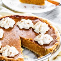 pumpkin pie topped with whipped cream with a slice on a plate in the background.