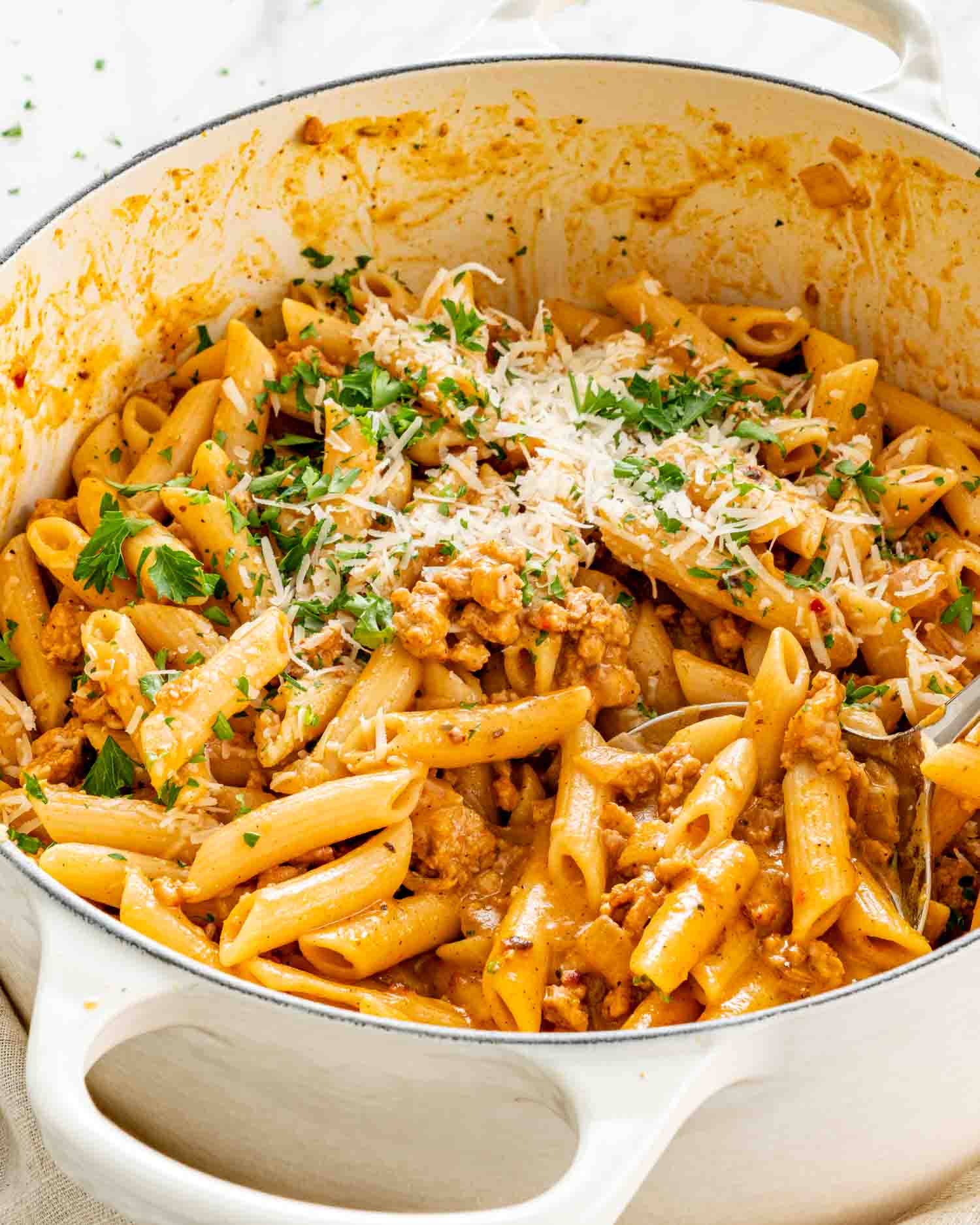 A serving spoon holds creamy pumpkin sausage penne above a white Dutch oven, with Parmesan and parsley scattered over the pasta.