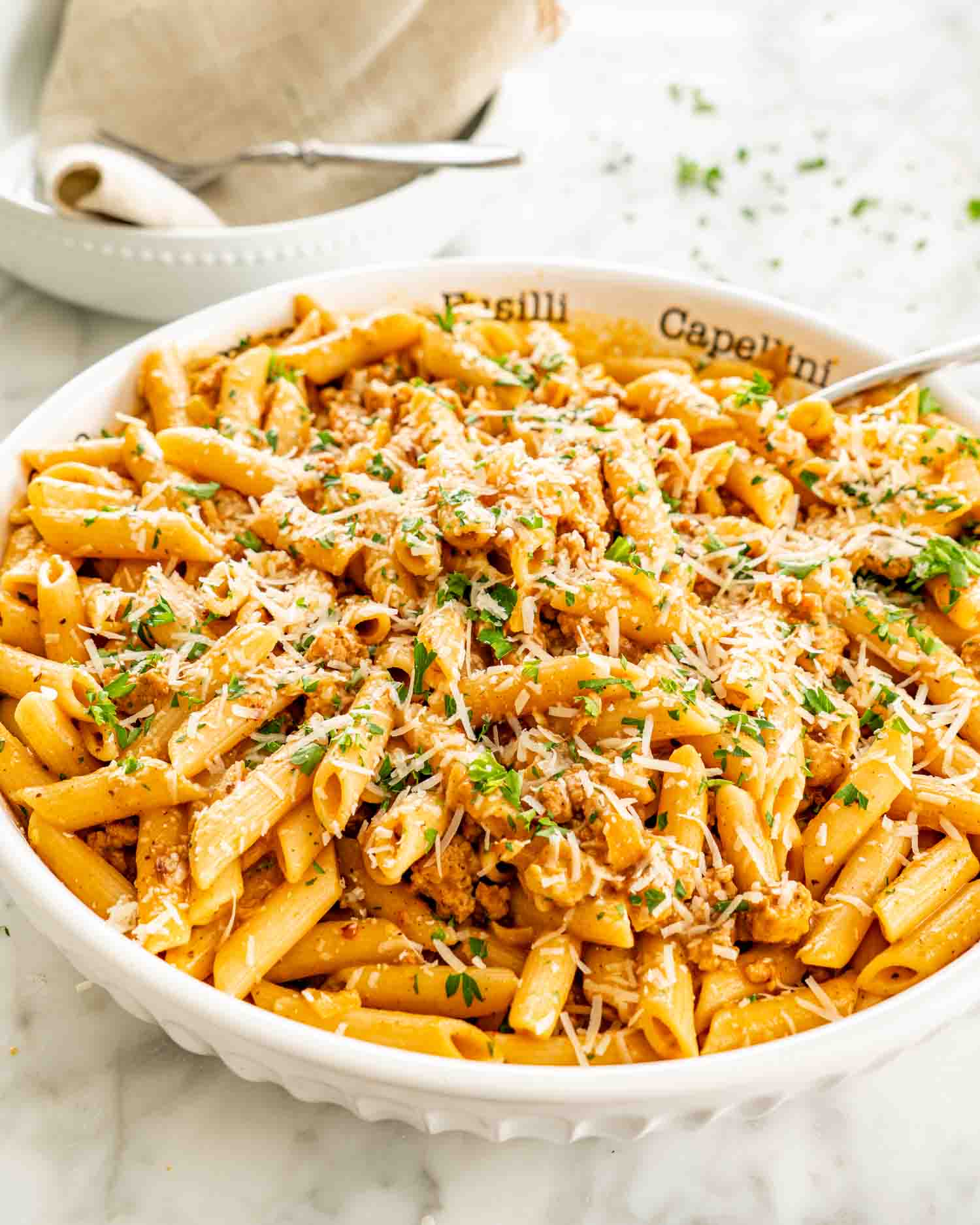 White serving bowl of creamy pumpkin sausage pasta with Parmesan and parsley, surrounded by white dishes and a beige napkin.