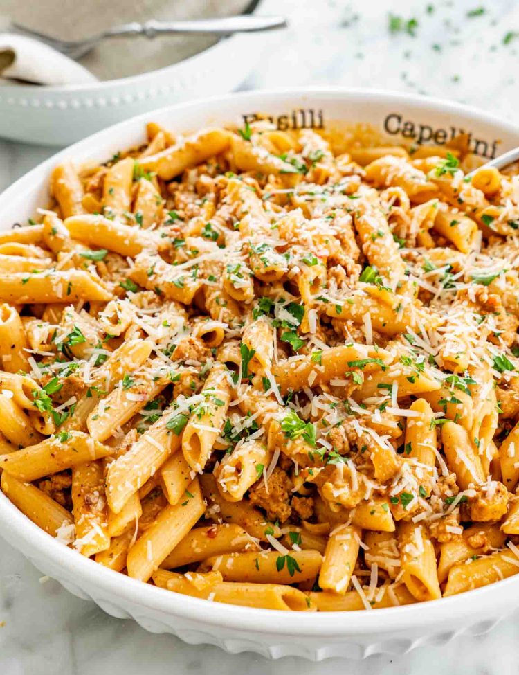Pumpkin sausage penne topped with Parmesan and parsley in a white serving bowl, with a spoon and stacked dishes nearby.