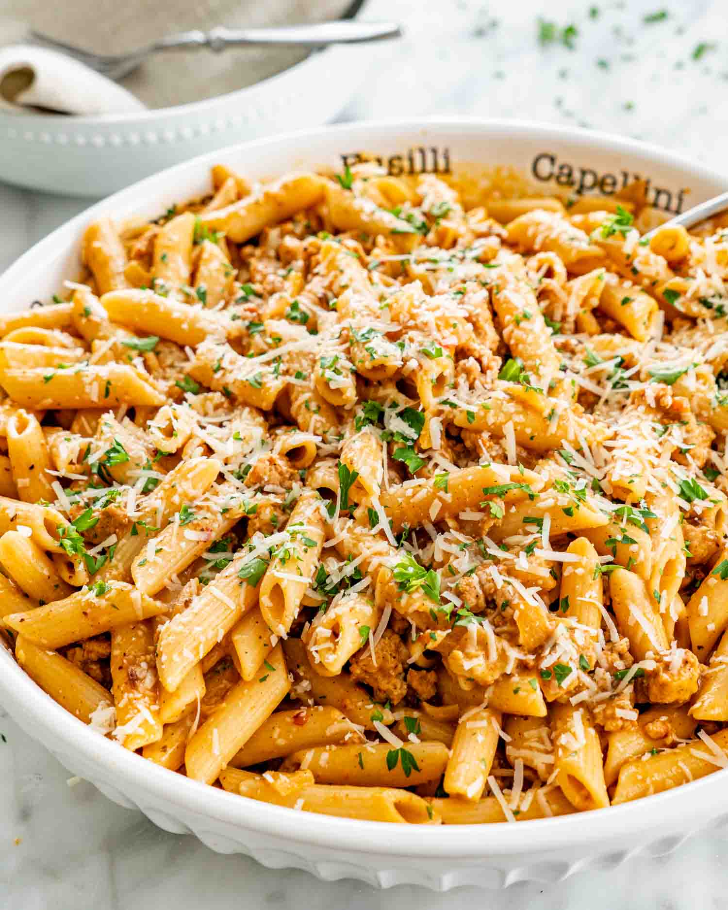 Pumpkin sausage penne topped with Parmesan and parsley in a white serving bowl, with a spoon and stacked dishes nearby.