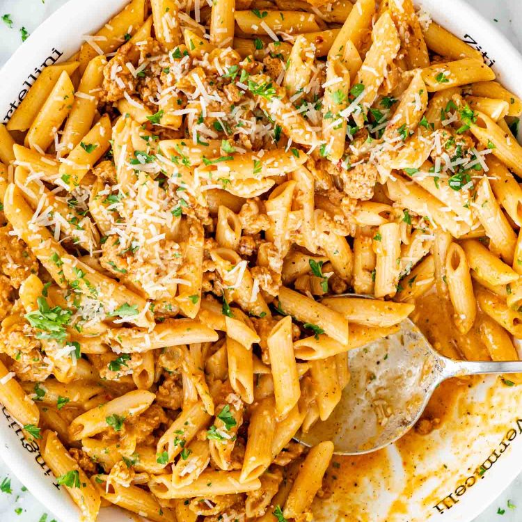Partially served pumpkin sausage pasta with Parmesan and parsley in a white bowl, with a silver spoon resting inside.