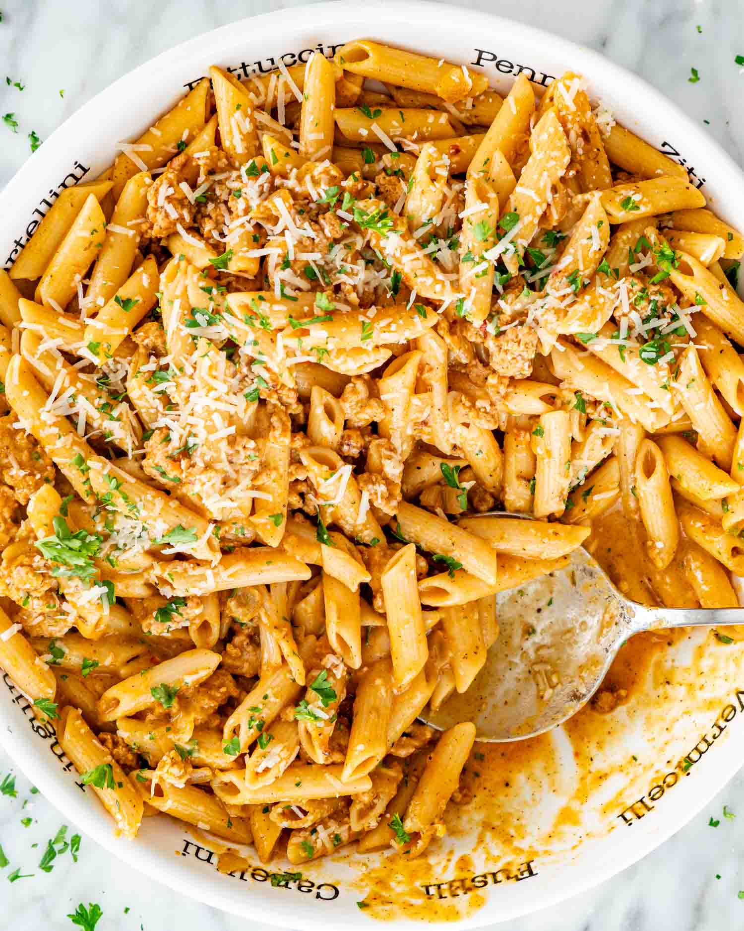 Partially served pumpkin sausage pasta with Parmesan and parsley in a white bowl, with a silver spoon resting inside.