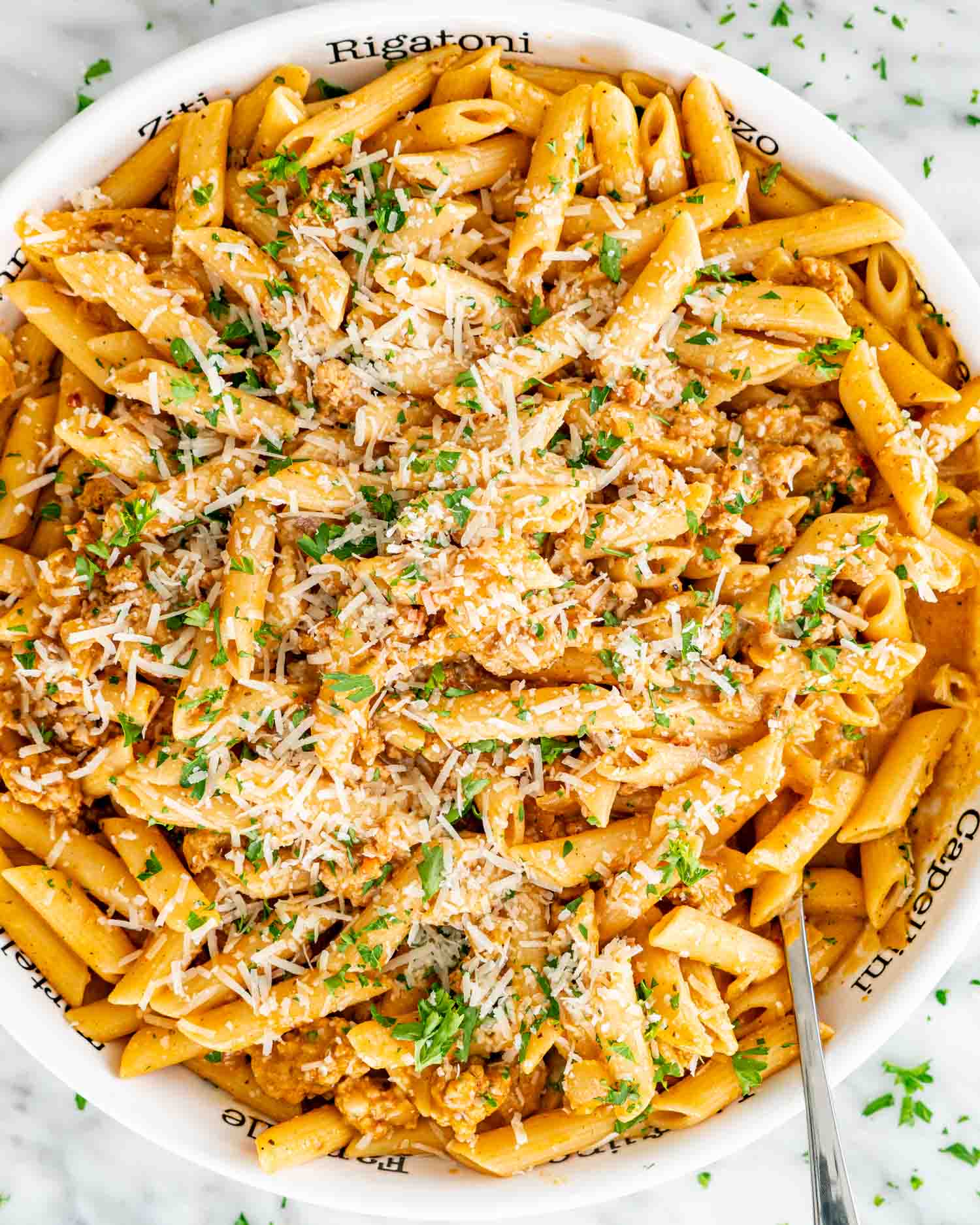 Overhead view of a white serving bowl filled with pumpkin sausage penne, topped with grated Parmesan and chopped parsley.