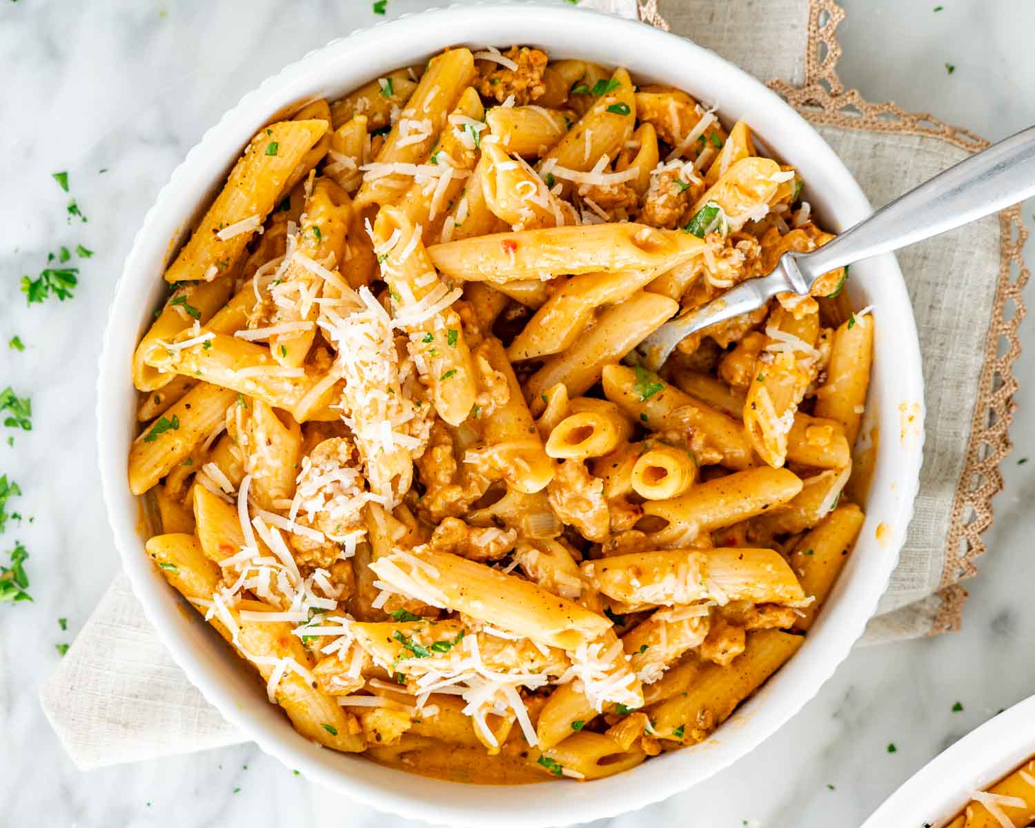 Overhead view of creamy pumpkin sausage pasta topped with Parmesan and parsley, beside a fork on a beige napkin.