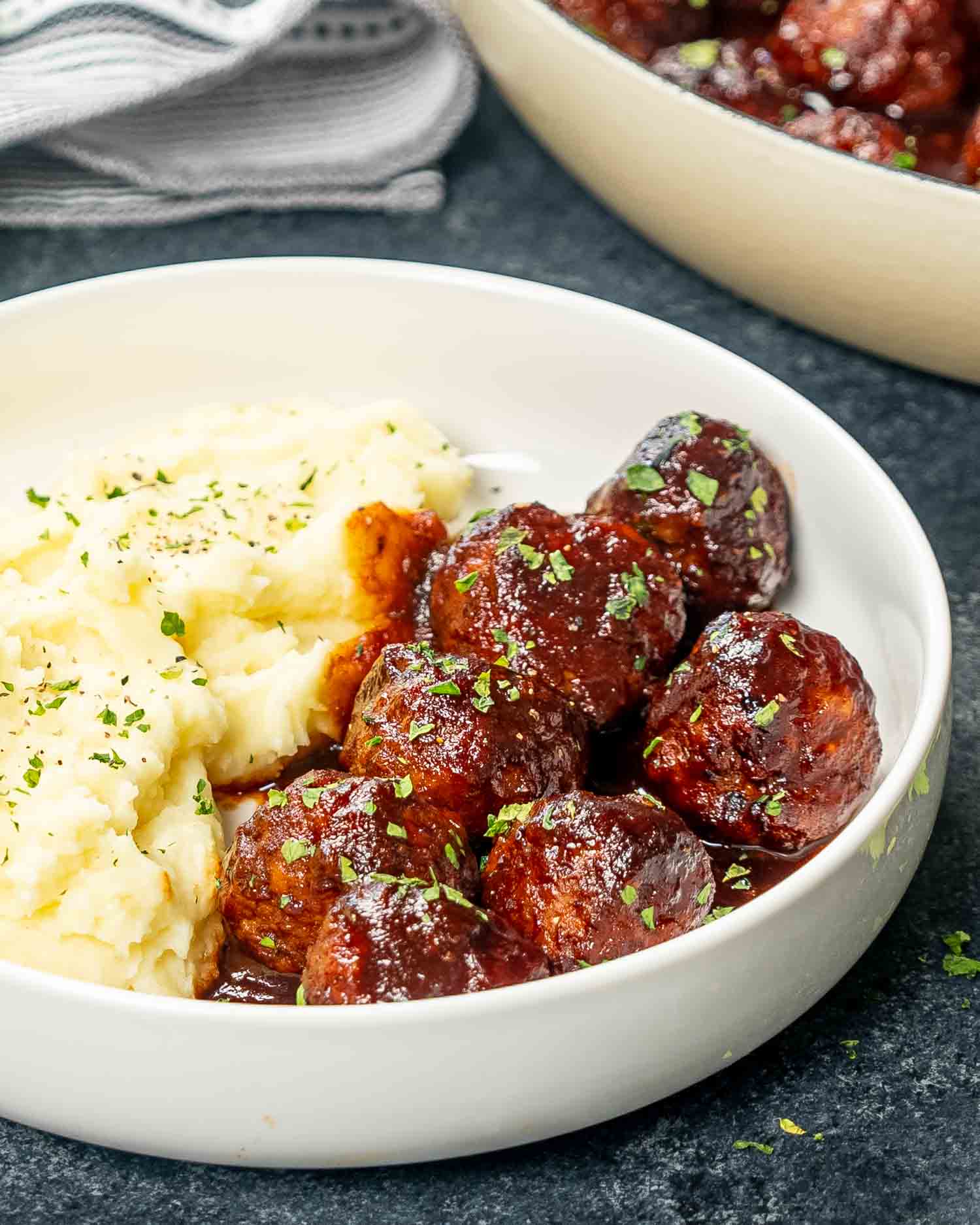 A delicious serving of stout meatballs, glazed with a smoky BBQ sauce, presented next to a creamy bowl of mashed potatoes in a white dish, showcasing the perfect comfort food combination.