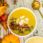 butternut squash soup in a white bowl garnished with pepitas, bacon, goat cheese and maple syrup.