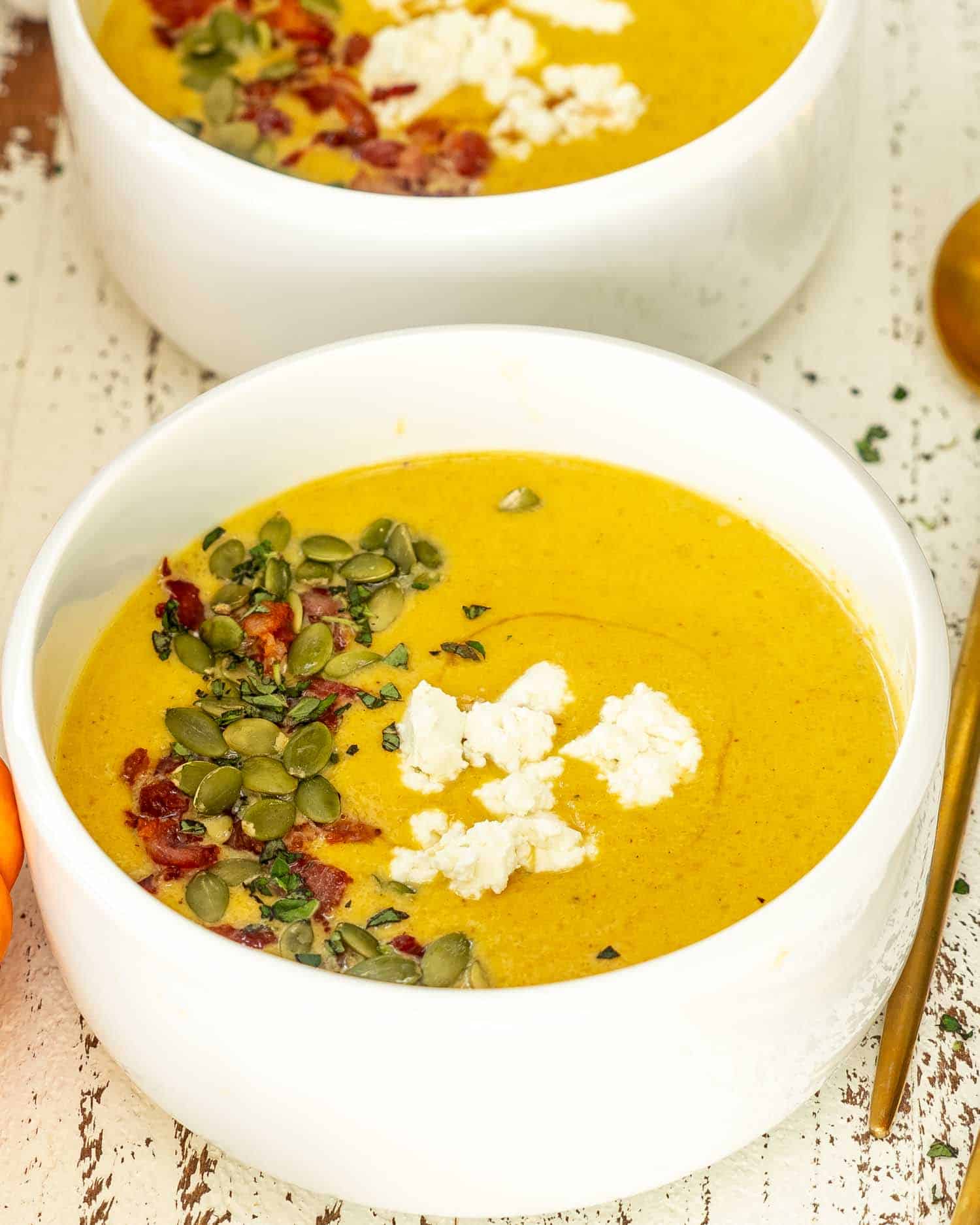 butternut squash soup in a white bowl garnished with pepitas, bacon, goat cheese and maple syrup.
