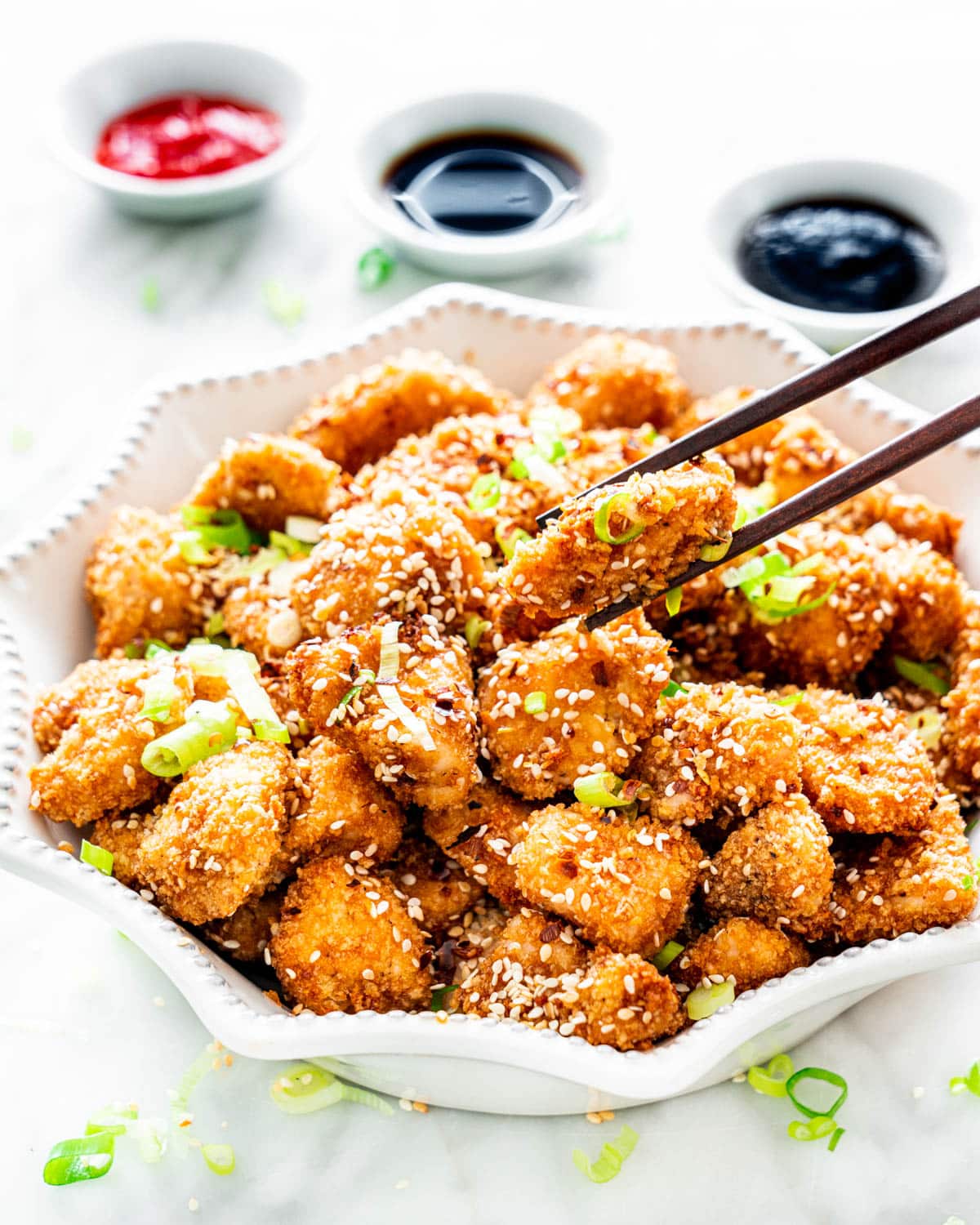 a white bowl loaded with honey sesame chicken with 3 sauces in the background