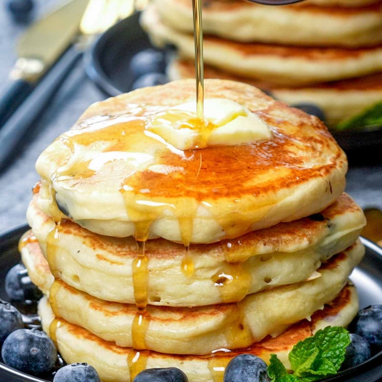 drizzling of maple syrup over a stack of lemon blueberry ricotta pancakes.