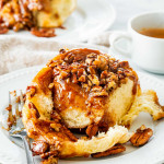 a gorgeous sticky bun on a white plate with a fork and a cup of espresso in the background.