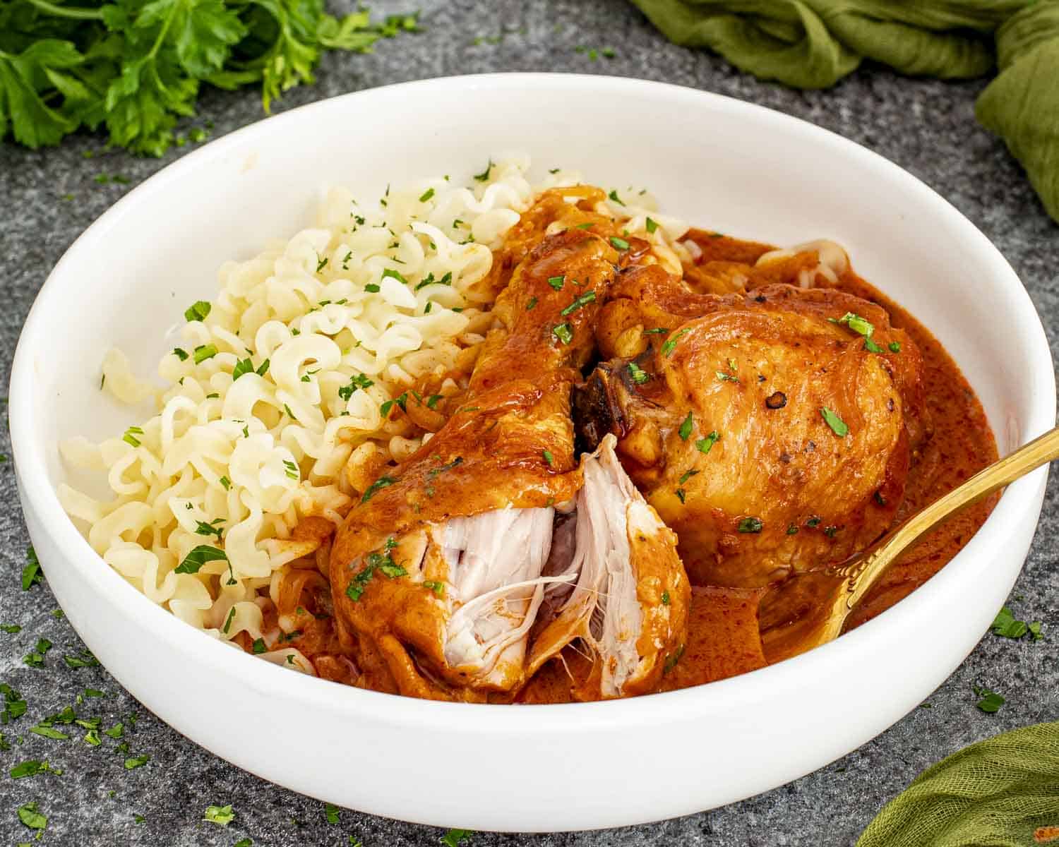 Close-up of chicken paprikash showing juicy chicken pulled apart in creamy paprika sauce with noodles.