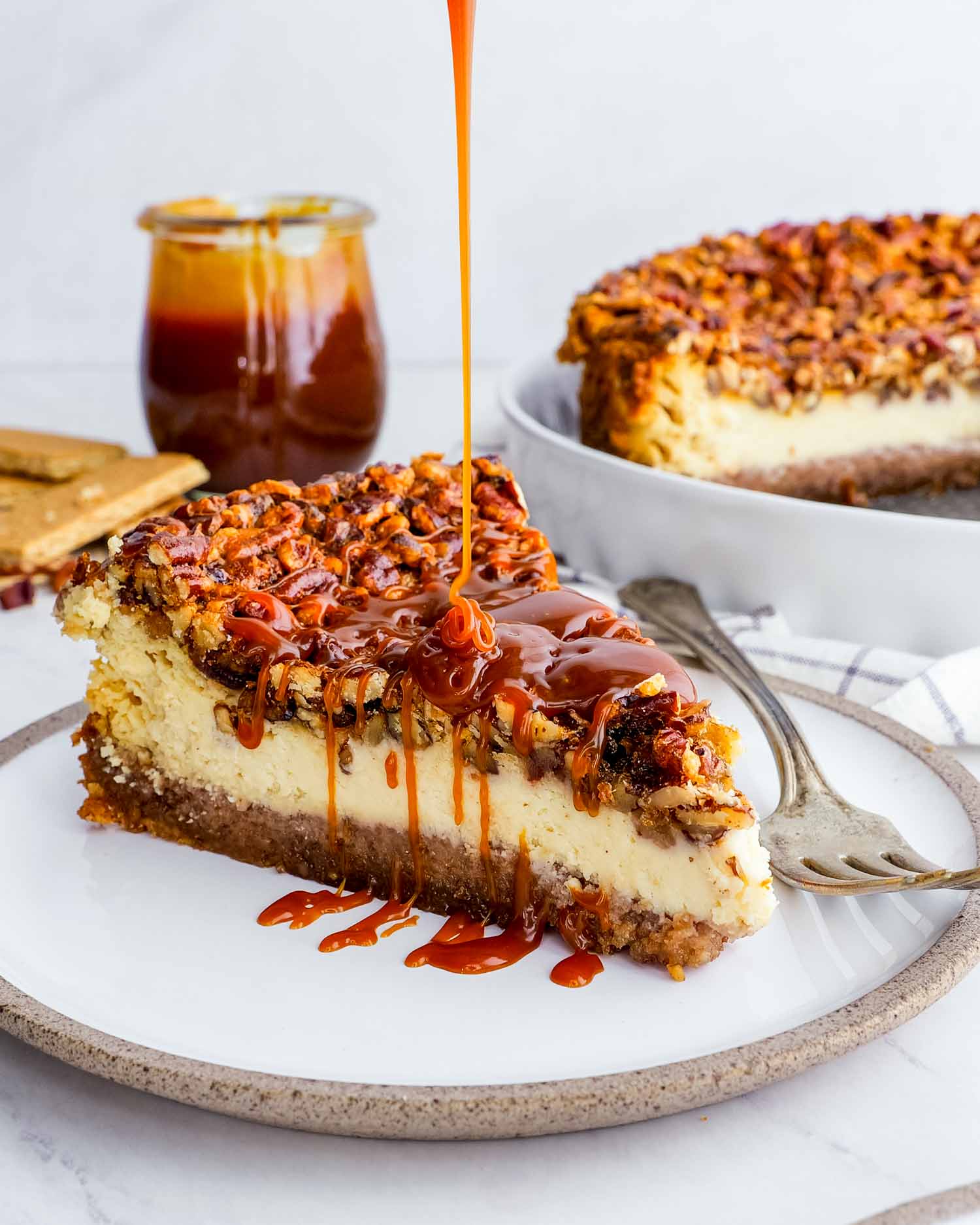 a slice of pecan pie cheesecake drizzled with caramel topping on a white plate.
