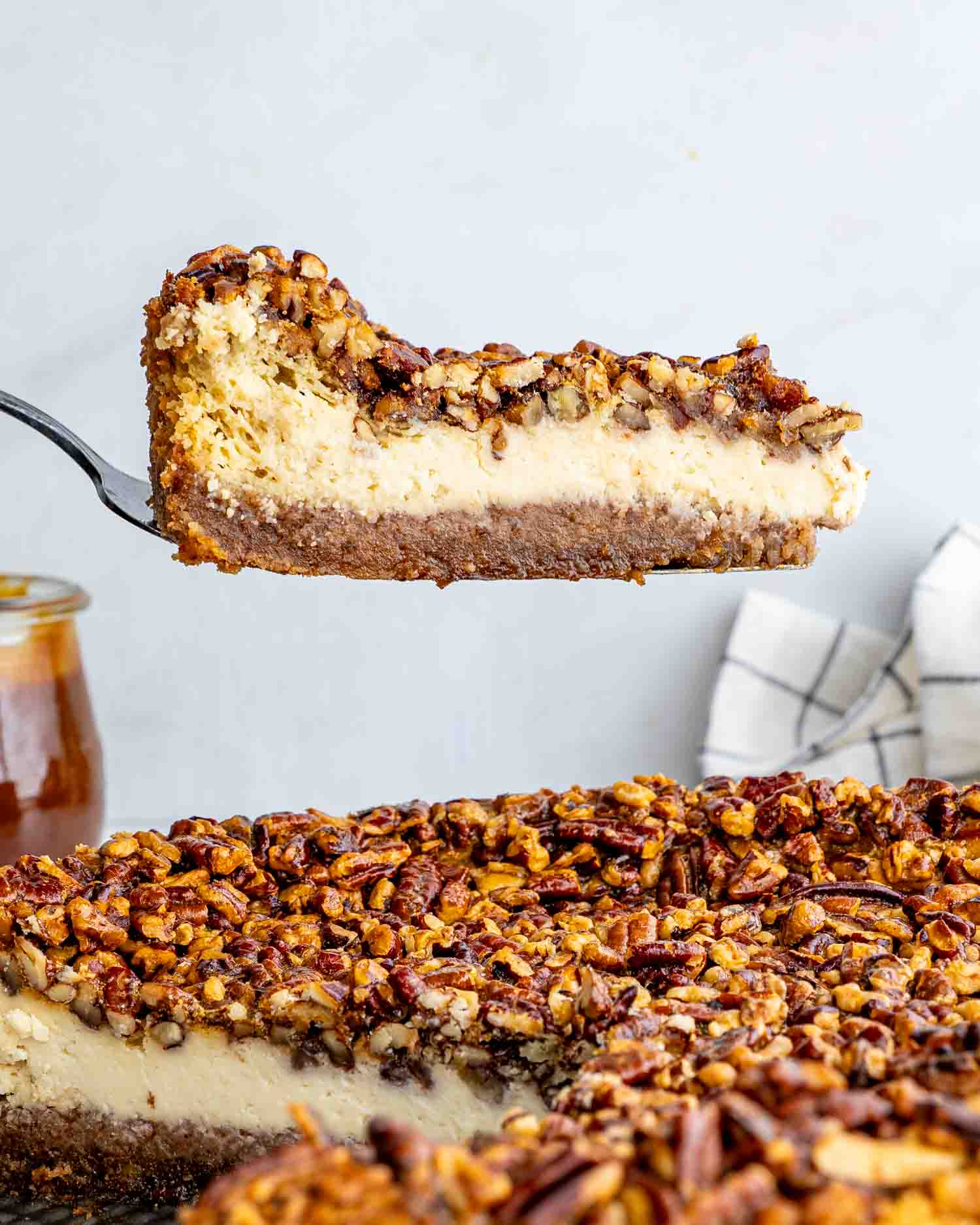 A delectable slice of Pecan Pie Cheesecake being lifted by a spatula, showcasing its creamy cheesecake layer and crunchy pecan topping, set against the backdrop of the whole cheesecake.