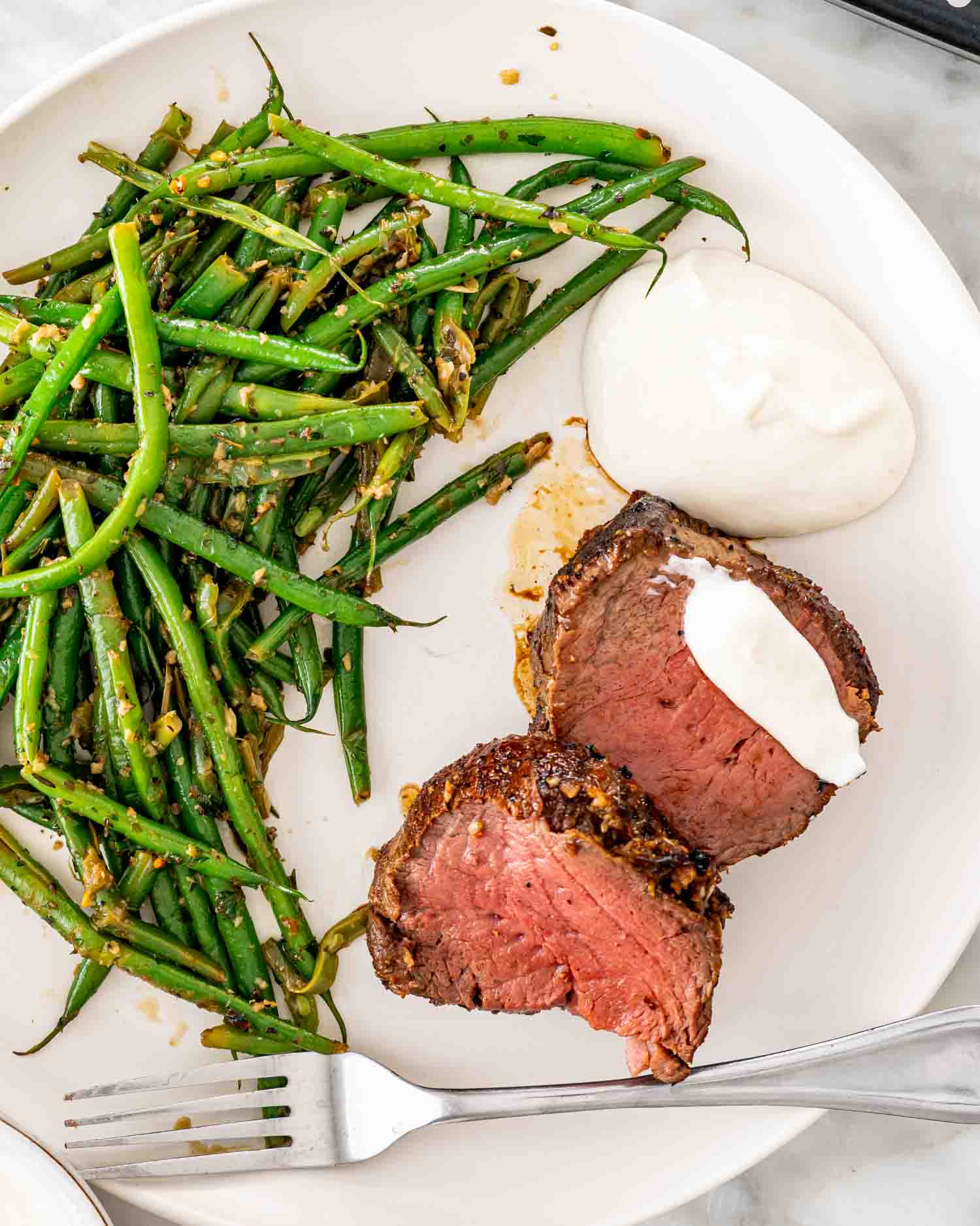2 slices of beef tenderloin with skillet green beans and horseradish yogurt sauce on a white plate.