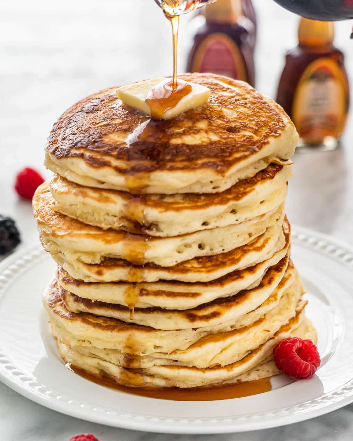 syrup being poured over a stack of buttermilk pancakes with a slab of butter on top