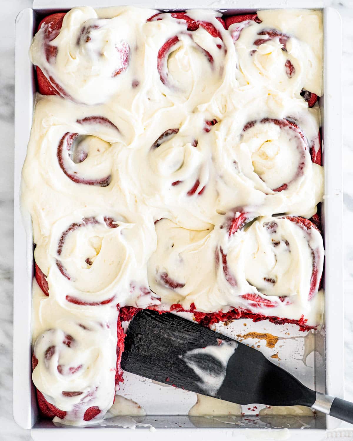 overhead shot of red velvet cinnamon rolls in a baking pan iced with cream cheese icing with 2 rolls missing