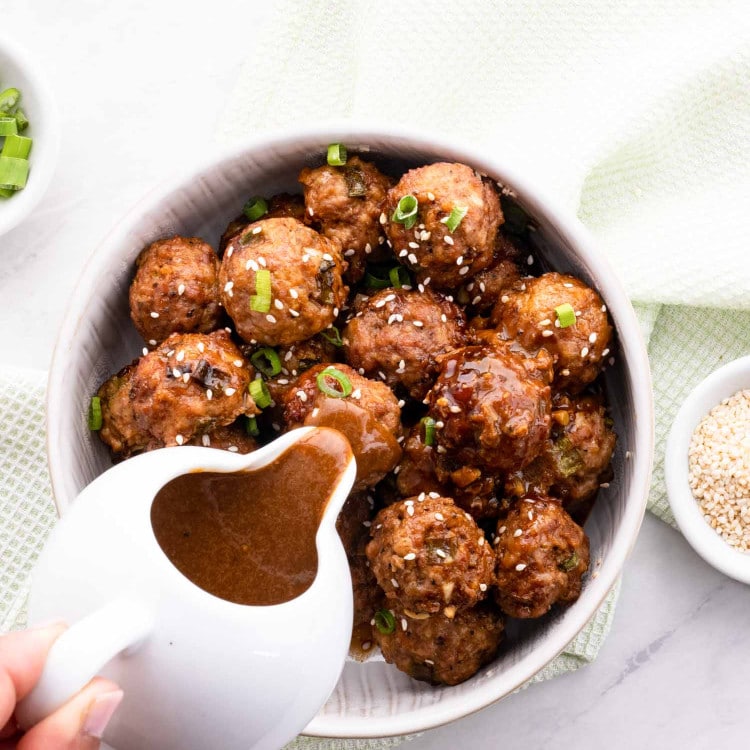 a hand holding a jug pouring an asian sauce over asian meatballs.