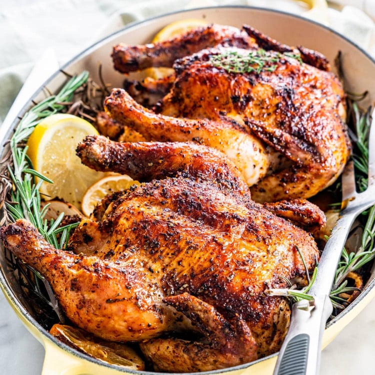 two roasted cornish hens in a roasting pan with sprigs of rosemary and lemon slices