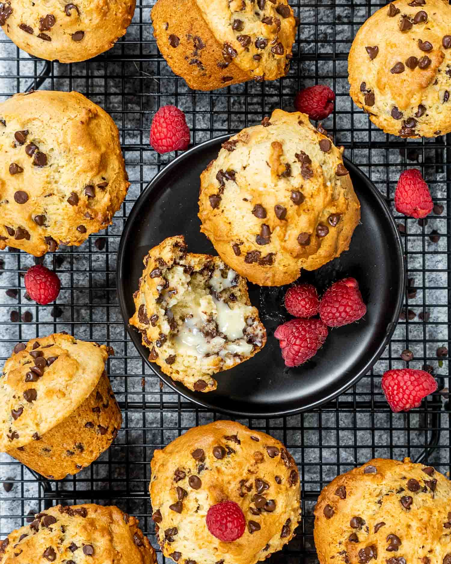 2 muffins, one with butter on a black plate surrounded by some other muffins and raspberries.